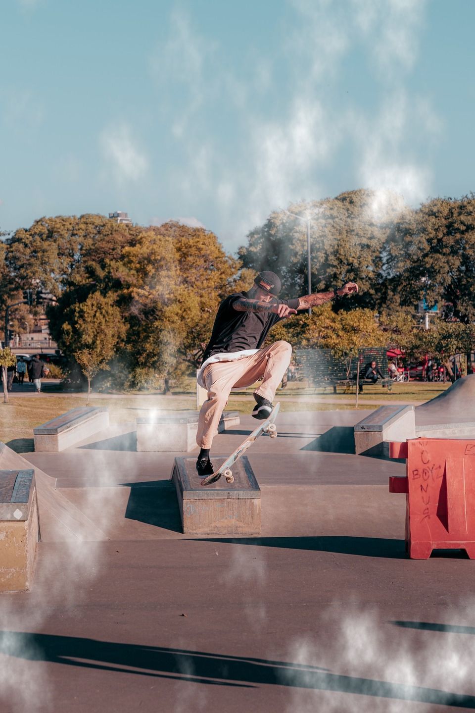 Cloud pattern style image of a person riding a skateboard while performing an aerial trick over a concrete obstacle at an outdoor skatepark, surrounded by ramps, rails, and trees.