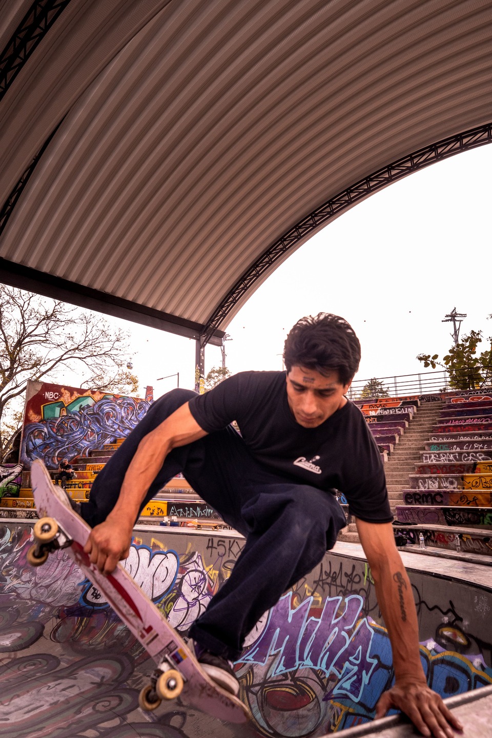 Skateboarder performing a trick inside a concrete skate bowl covered in colorful graffiti beneath a large curved canopy, with tiered seating and urban artwork visible in the background.
    