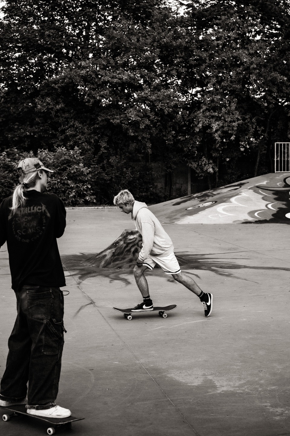 Black-and-white photo of two skateboarders at an outdoor skatepark. One skateboarder pushes across the concrete surface while another stands on a skateboard nearby, with graffiti-covered ramps and trees in the background.
