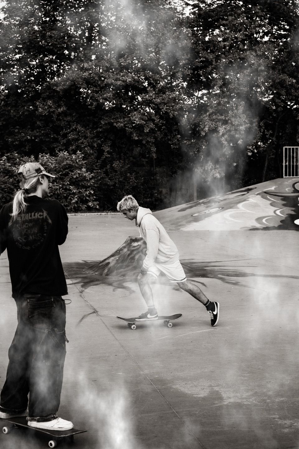 Cloud pattern style image of two skateboarders at an outdoor skatepark. One skateboarder pushes across the concrete surface while another stands on a skateboard nearby, with graffiti-covered ramps and trees in the background.